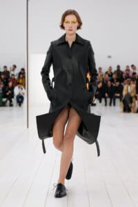 Model on a runway wearing a black leather-like coat with sculptural, winged hems and hands in pockets, audience in background.
