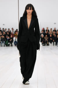 Model walking a fashion show in an oversized black suit with a plunging blazer and wide draped pants, white shoes, as an audience watches from the background.