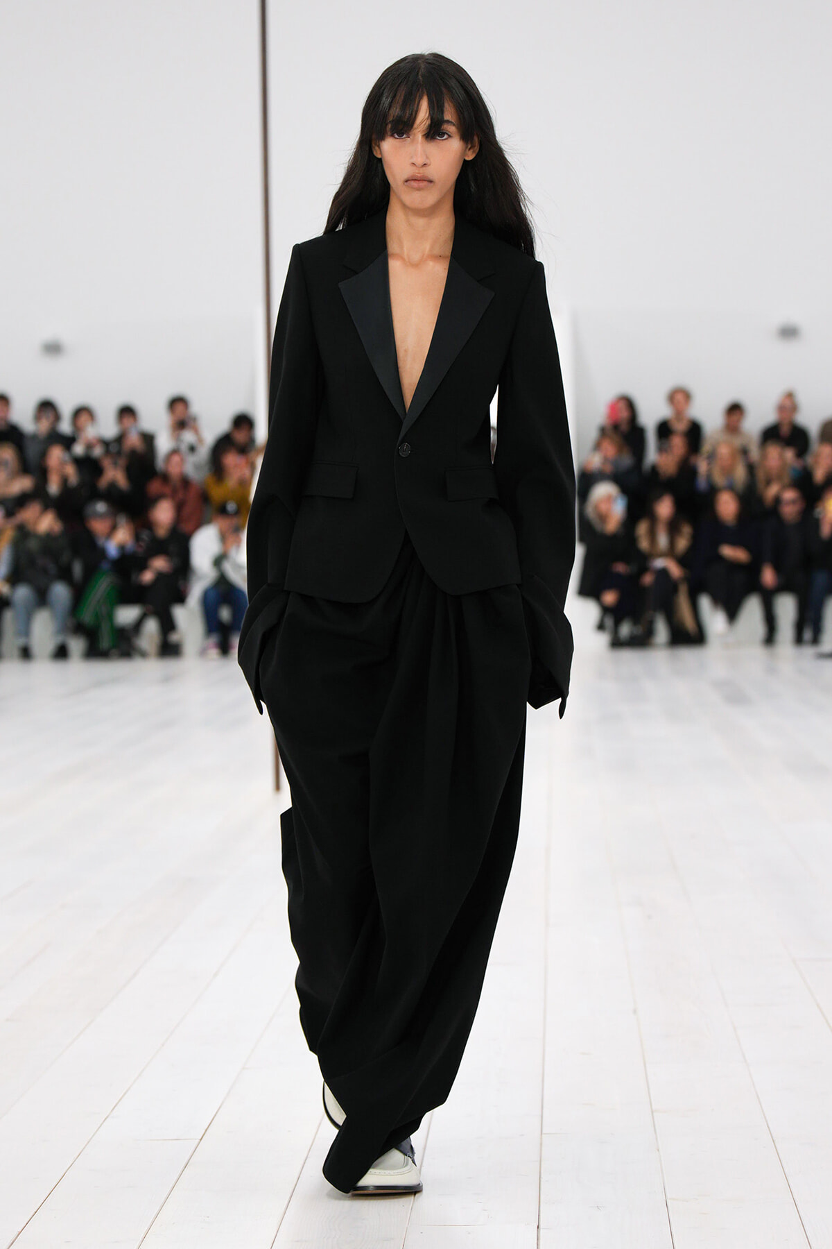 Model walking a fashion show in an oversized black suit with a plunging blazer and wide draped pants, white shoes, as an audience watches from the background.