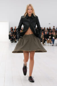 Model on a runway wearing a cropped black leather jacket and olive A-line skirt with a gold trim, walking toward the camera as an audience watches.