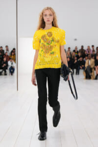 Model walking runway in a bright yellow floral top and black slim pants, holding a dark handbag as audience watches.