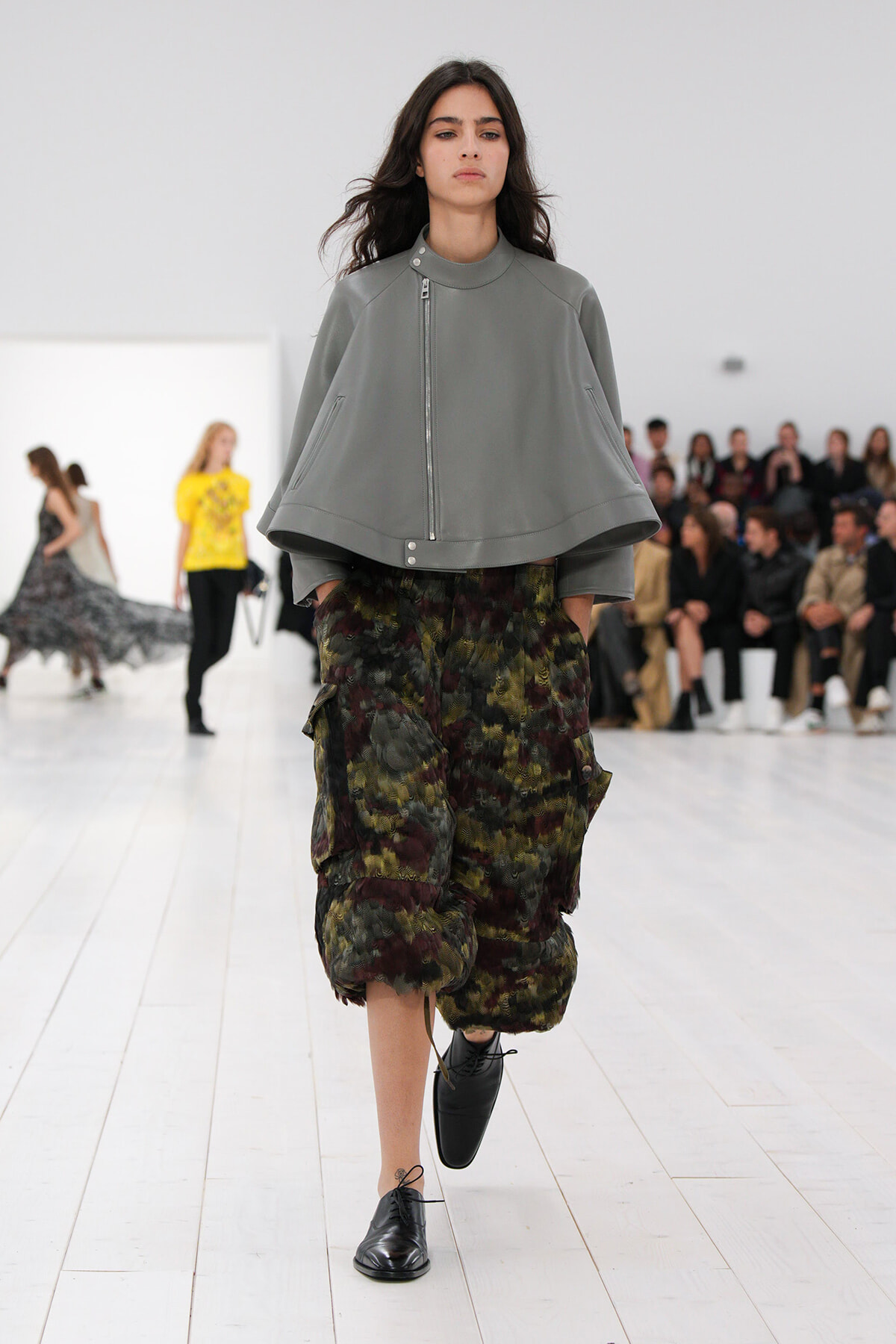 Model walking a runway in a gray zip-front cape over camouflage cargo pants; white floor and audience in the background.