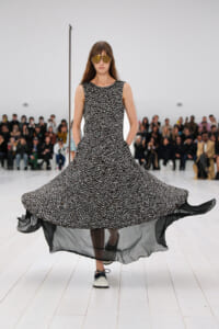 Model walks a fashion runway in a sleeveless black-and-white patterned dress with a flowing, sheer hem and oversized sunglasses.