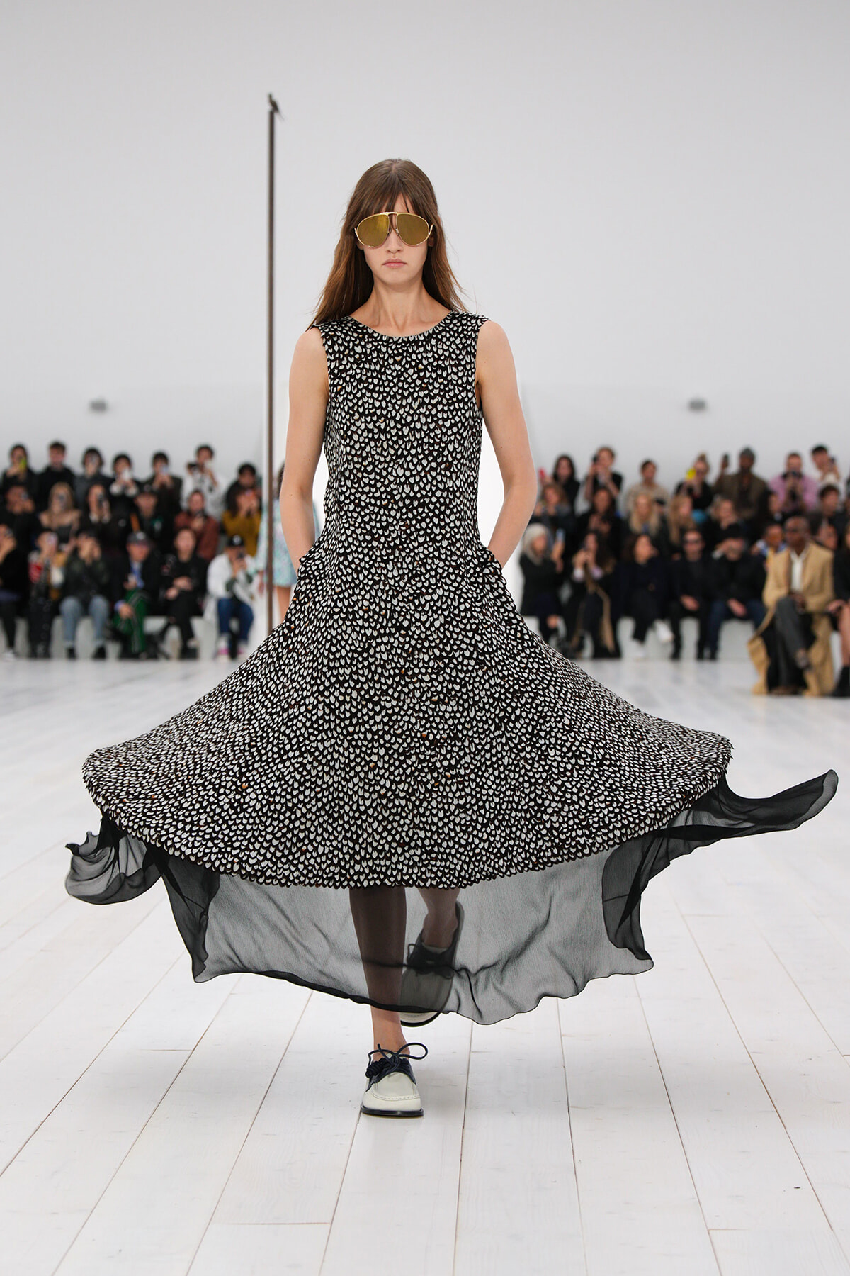 Model walks a fashion runway in a sleeveless black-and-white patterned dress with a flowing, sheer hem and oversized sunglasses.