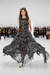 Model walking the runway in a sheer black floral gown with a flowing, layered skirt.