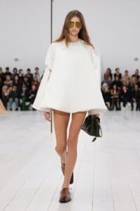 Model on a runway wearing a white textured, oversized dress with short sleeves, brown oxford shoes, and gold aviator sunglasses, carrying a black quilted handbag with a gold chain.