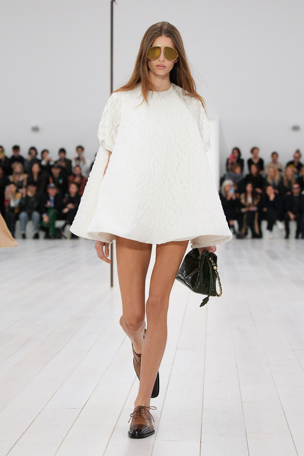 Model on a runway wearing a white textured, oversized dress with short sleeves, brown oxford shoes, and gold aviator sunglasses, carrying a black quilted handbag with a gold chain.