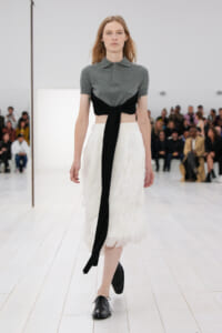 Model walking a runway in a gray short-sleeve polo with a black tie belt over a white feathered midi skirt, black shoes visible, audience in background.