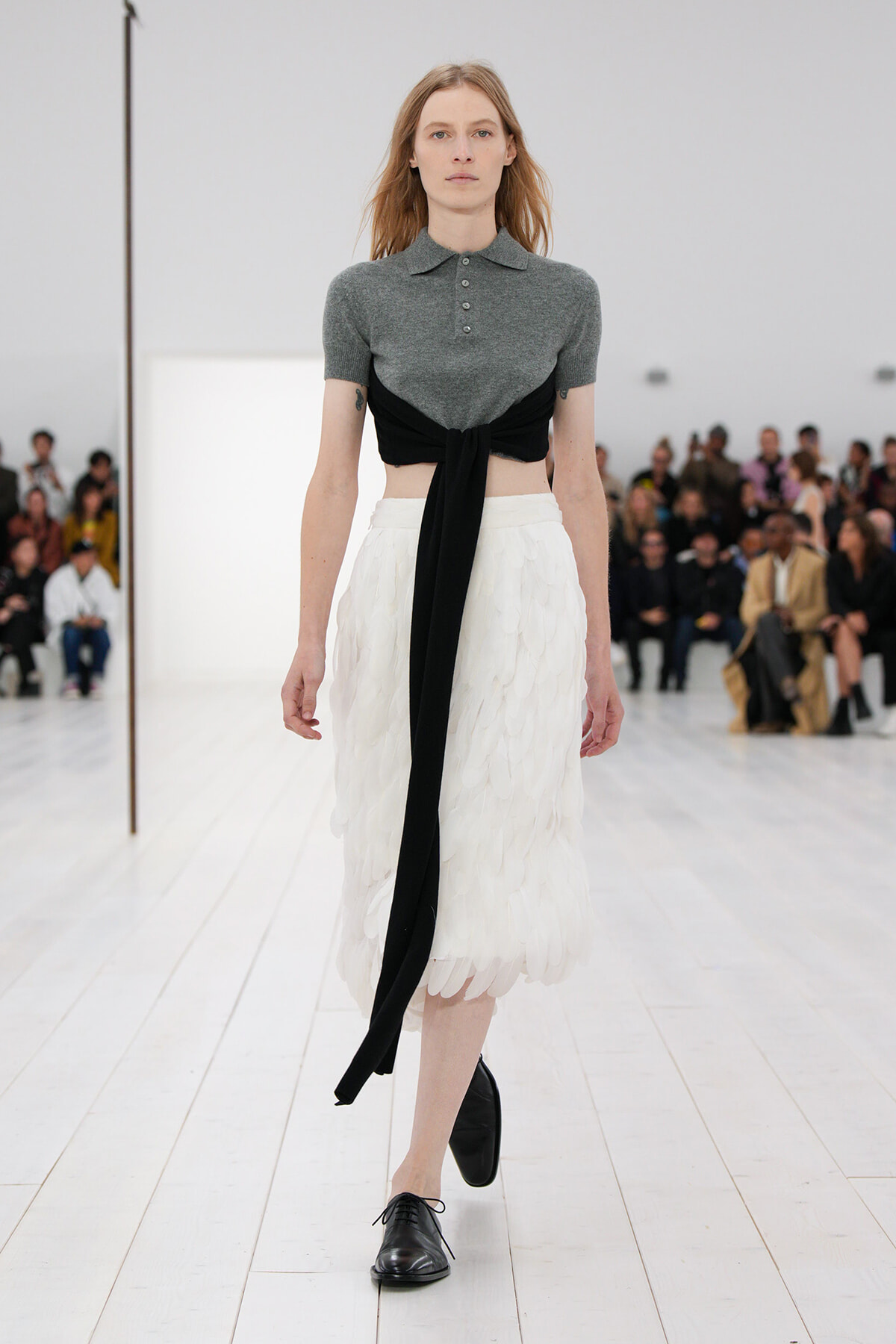 Model walking a runway in a gray short-sleeve polo with a black tie belt over a white feathered midi skirt, black shoes visible, audience in background.