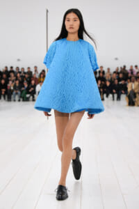 Model walking a runway in a bright blue textured oversized dress with black lace-up shoes; audience seated in the background.