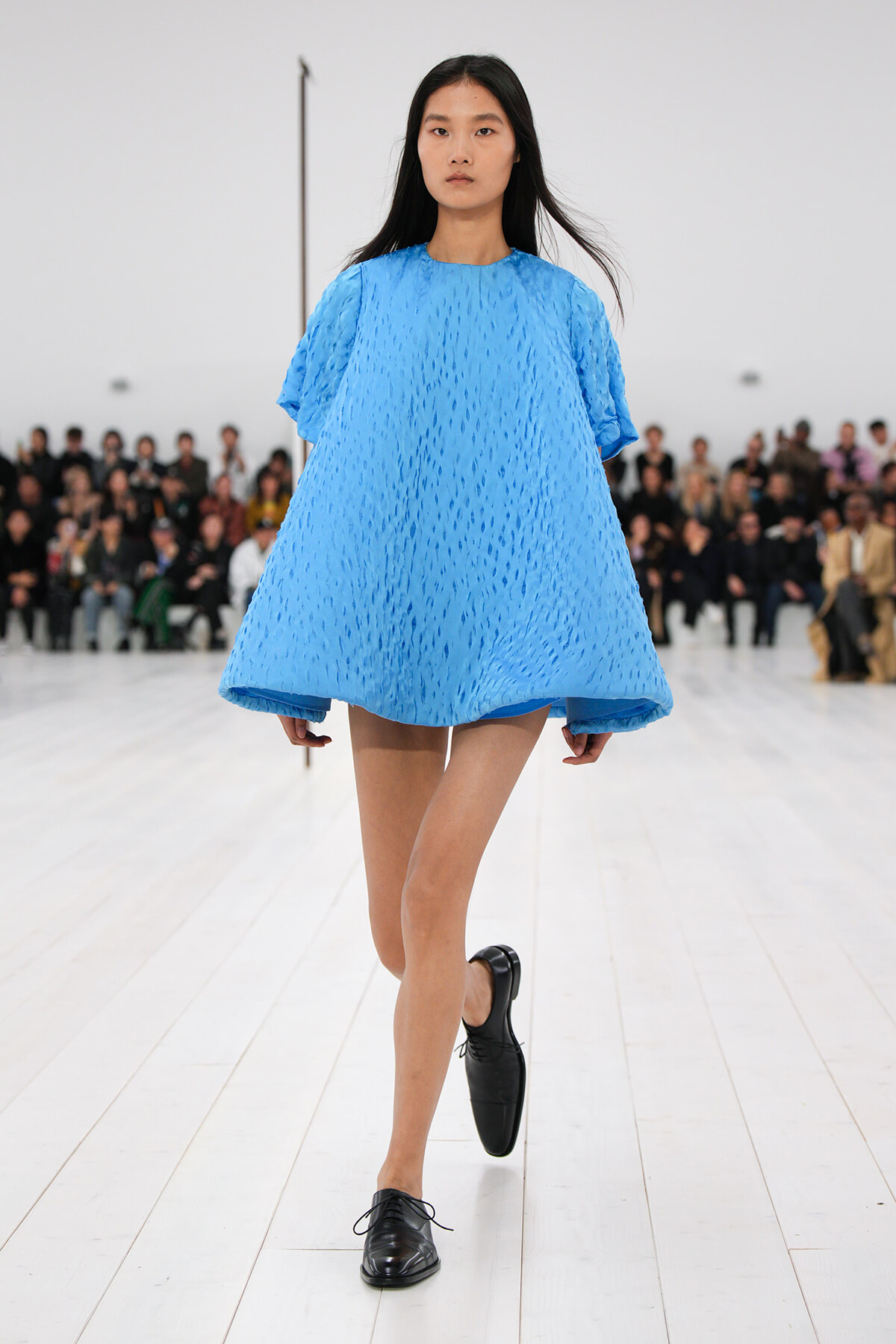Model walking a runway in a bright blue textured oversized dress with black lace-up shoes; audience seated in the background.
