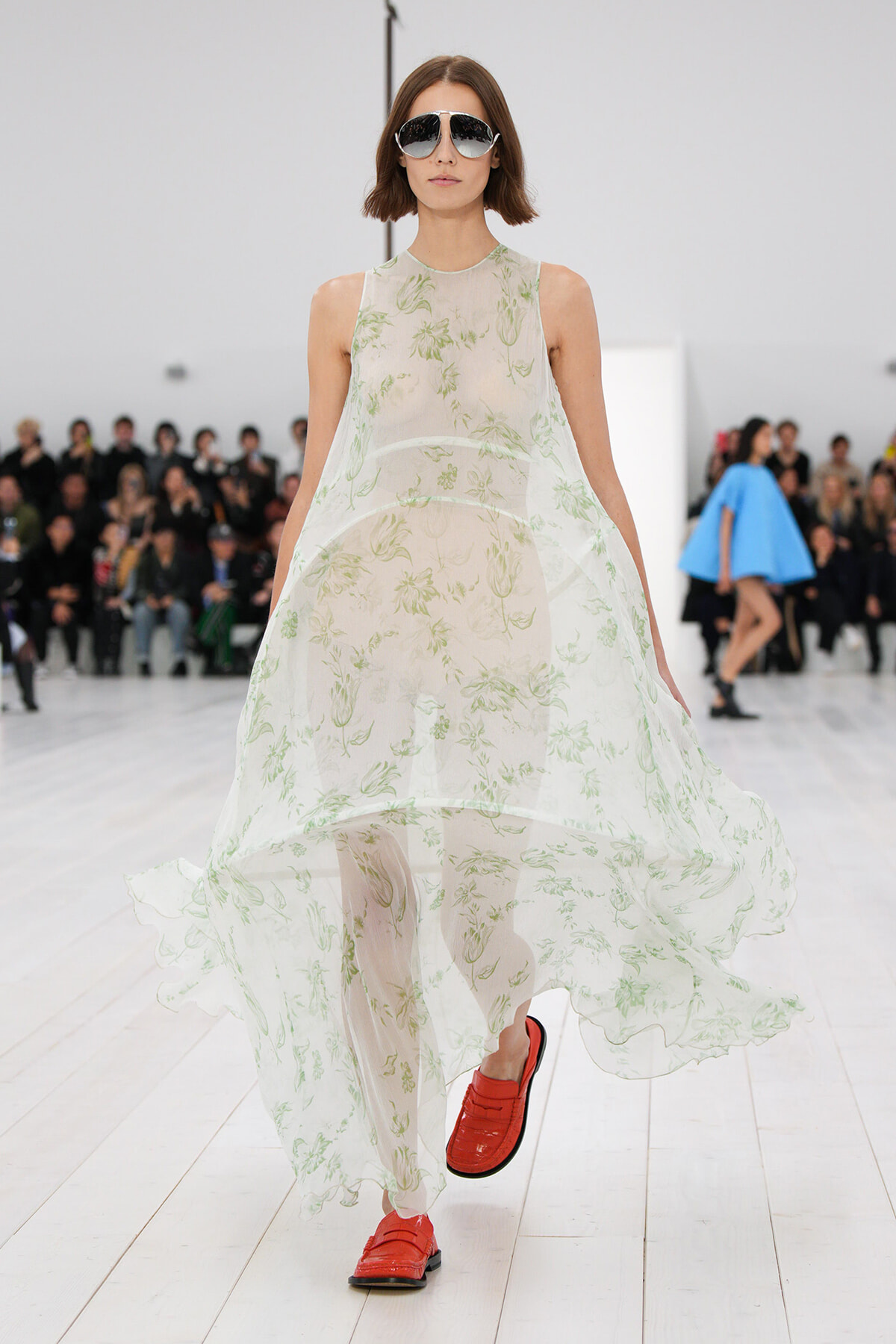 Runway model in a sheer white floral dress with green print, wearing oversized sunglasses and red loafers.