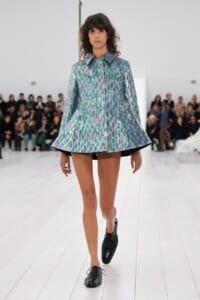 Model walks a fashion runway in a metallic iridescent cape-style coat with a structured collar, paired with black shorts and black lace-up shoes.