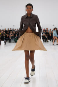 Model walks the runway in a cropped brown leather jacket paired with a beige A-line skirt and two-tone shoes, audience in the background.