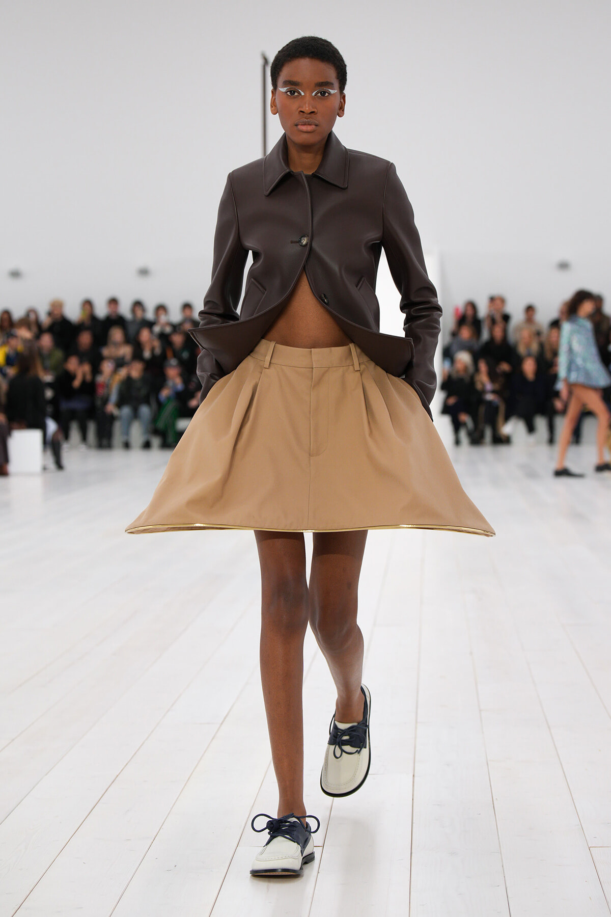 Model walks the runway in a cropped brown leather jacket paired with a beige A-line skirt and two-tone shoes, audience in the background.