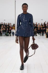 Model walking a fashion show runway in a navy sequin long-sleeve mini dress, carrying a burgundy handbag with a long strap behind her.