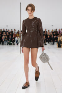 Model walking a runway in a brown sequin mini dress, brown two-tone oxford shoes, and a gray structured handbag by her side.