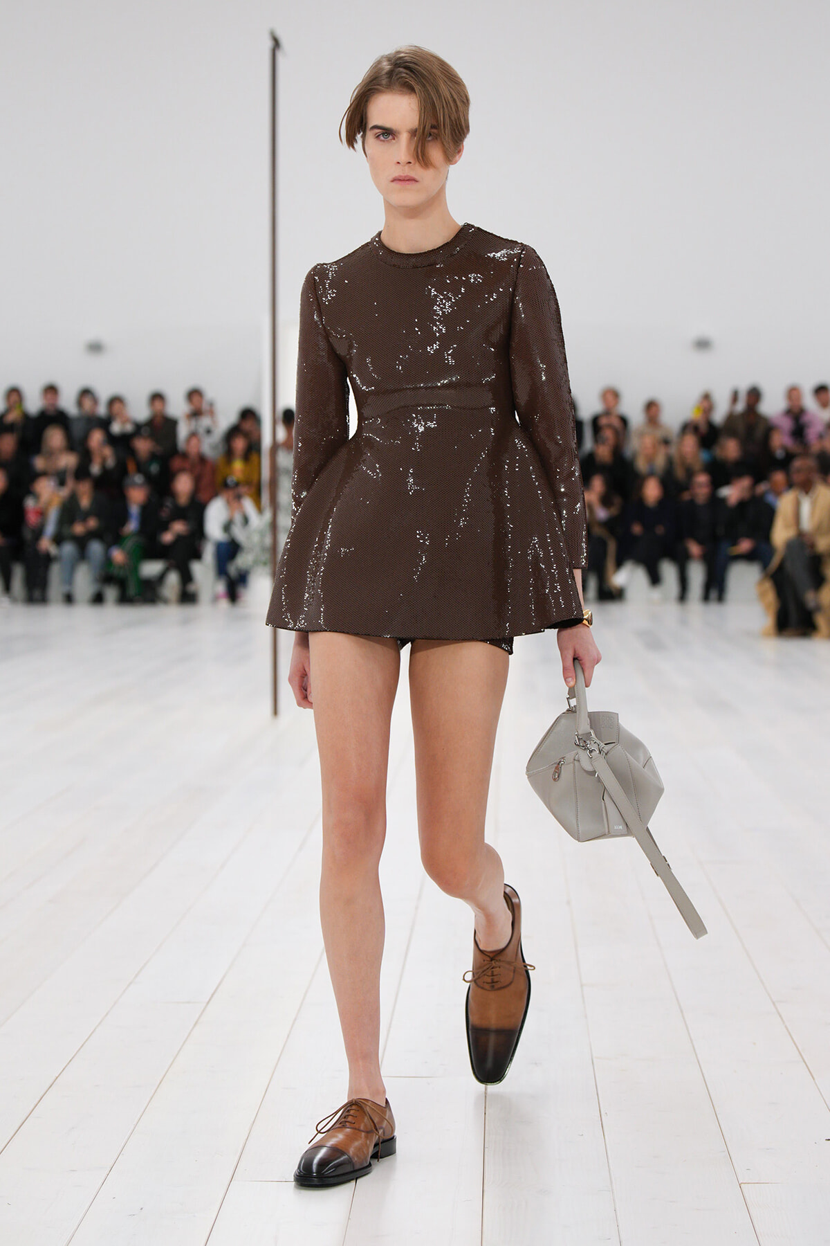 Model walking a runway in a brown sequin mini dress, brown two-tone oxford shoes, and a gray structured handbag by her side.