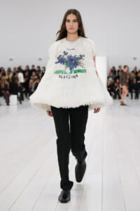 Female model walks a fashion runway in a voluminous white feathered top with a blue floral print, paired with black tailored pants and black shoes.