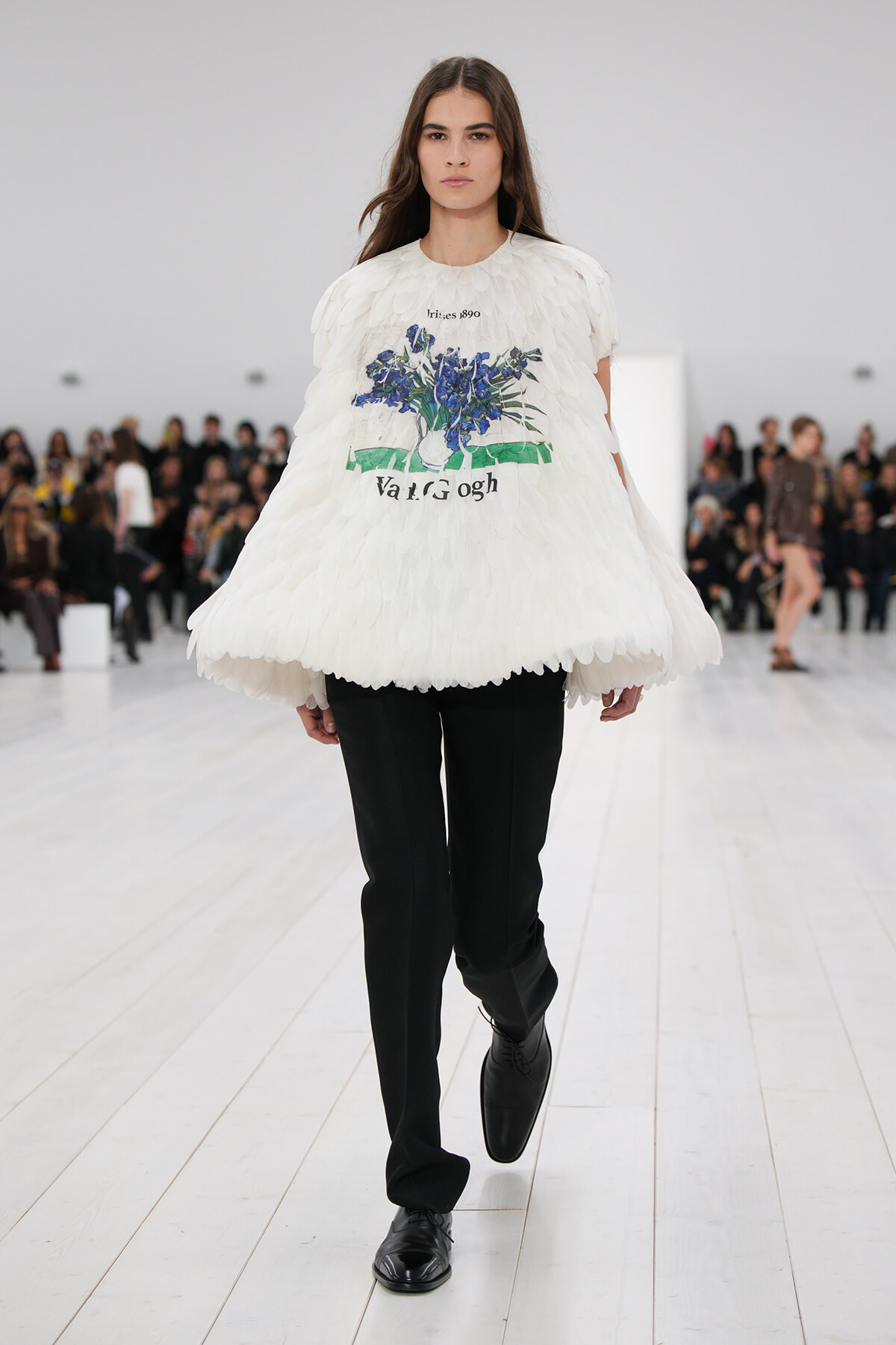 Female model walks a fashion runway in a voluminous white feathered top with a blue floral print, paired with black tailored pants and black shoes.