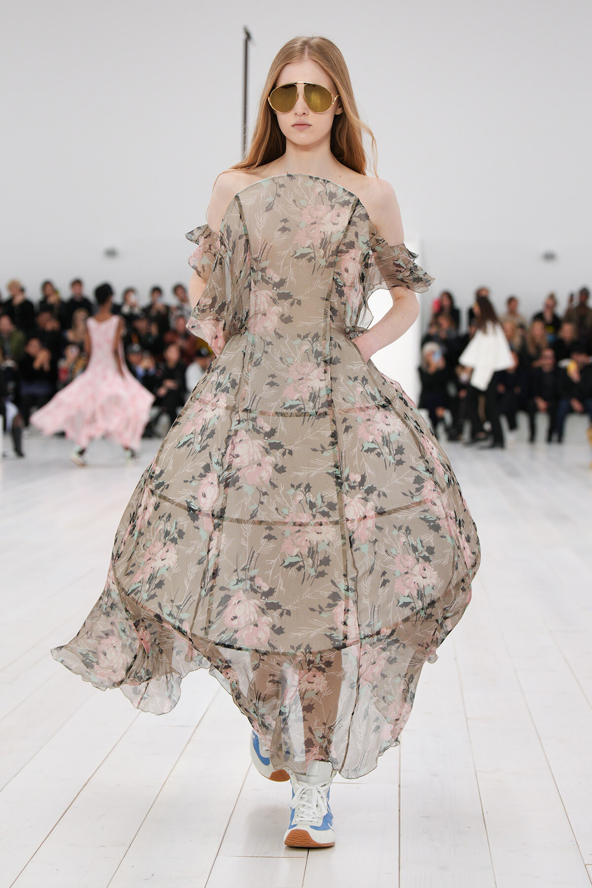 Model walking a runway in a beige floral off-shoulder chiffon gown with pink roses, large sunglasses, and blue-and-white sneakers.