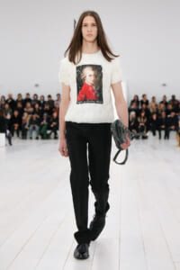 Model walking a fashion runway in a white textured T-shirt featuring a portrait graphic, paired with black trousers and black shoes, carrying a gray handbag.