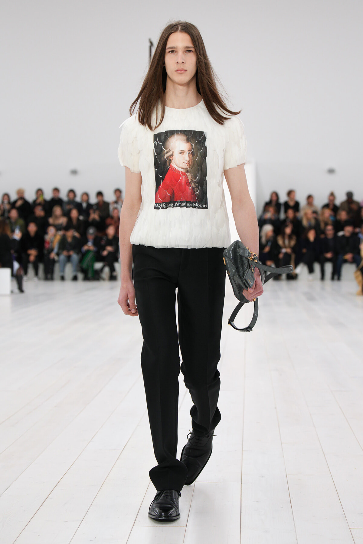 Model walking a fashion runway in a white textured T-shirt featuring a portrait graphic, paired with black trousers and black shoes, carrying a gray handbag.