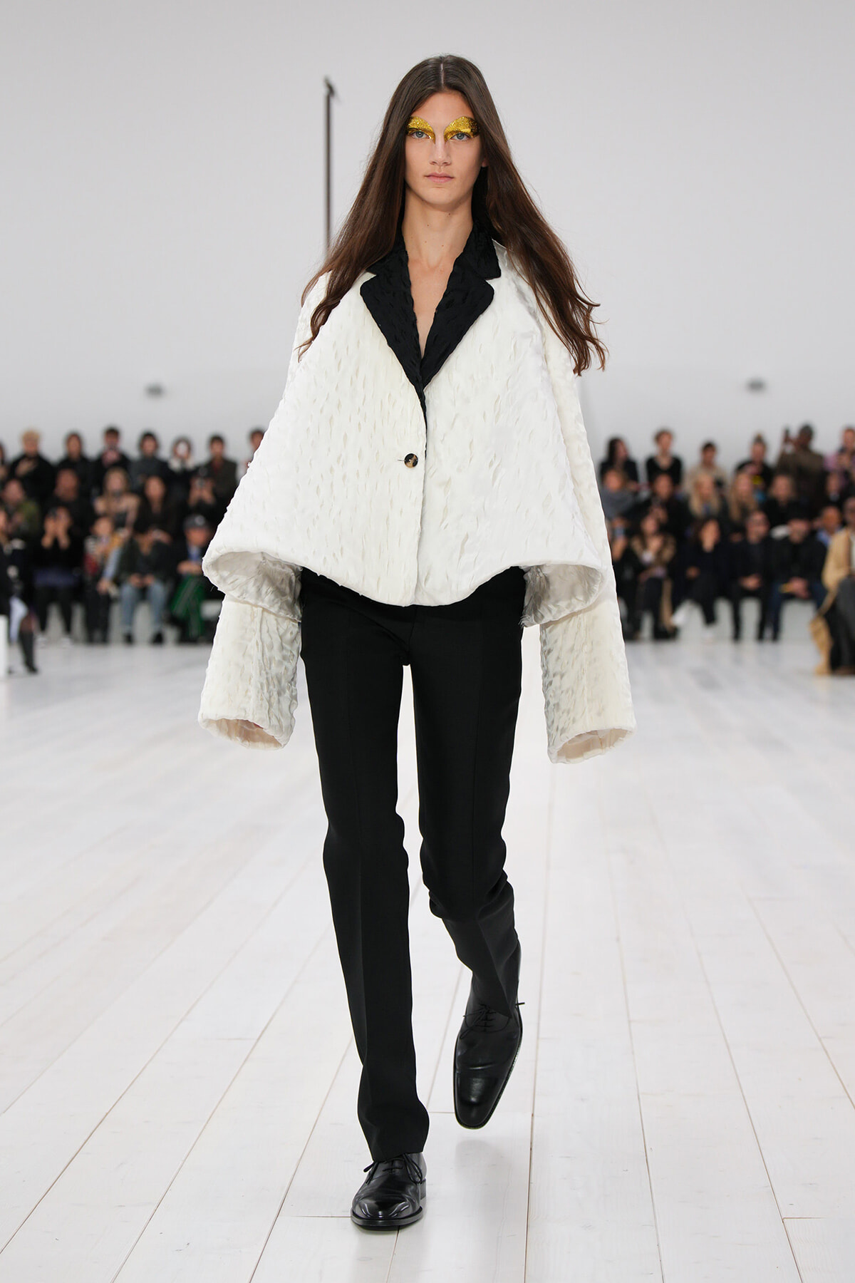 Female model walks a fashion show wearing a white textured cape coat with black lapels, black pants, and gold glitter eye makeup.