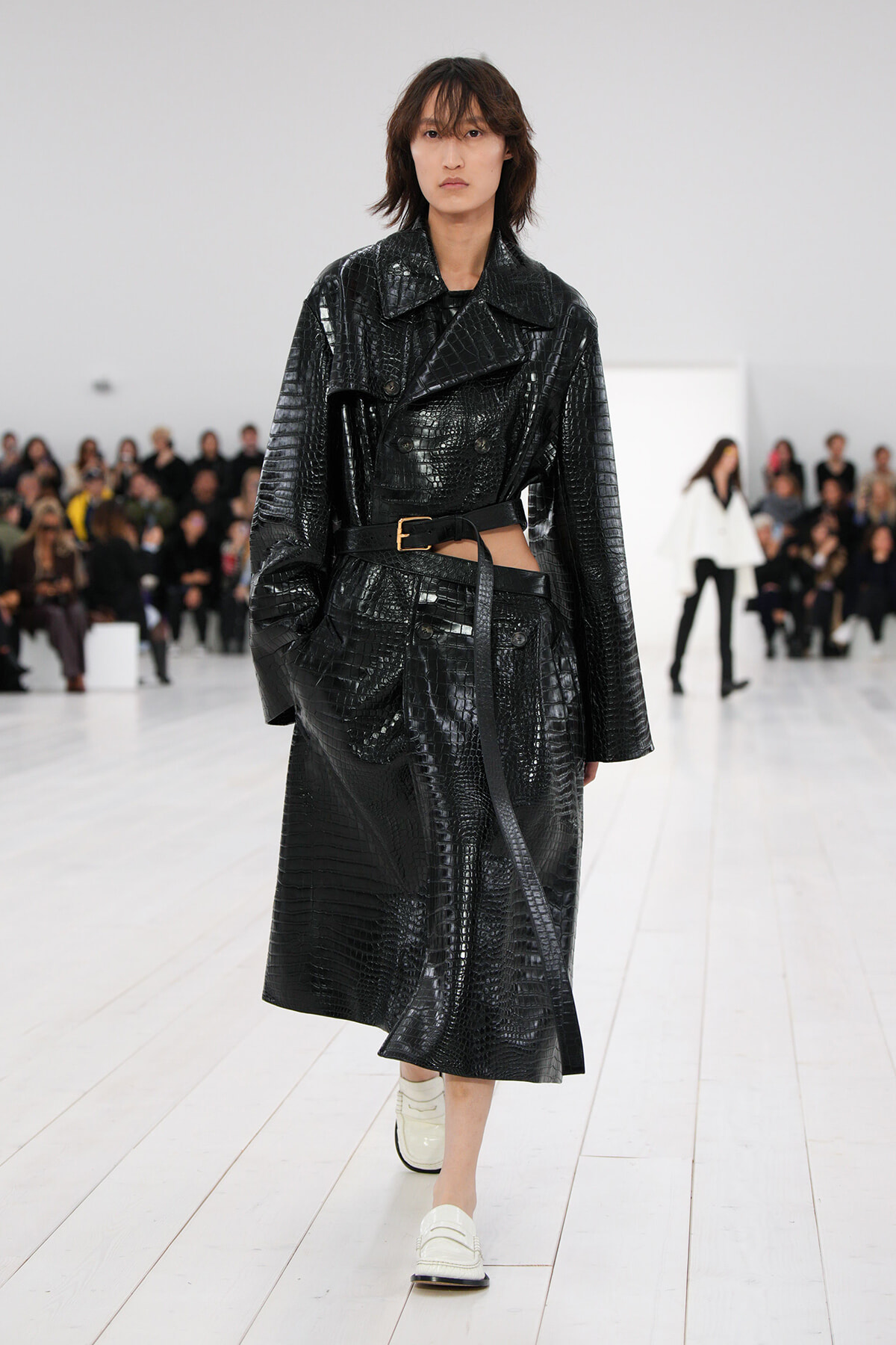 Model walking the runway in a black crocodile-embossed leather trench with a belt, white loafers, and an audience in the background.