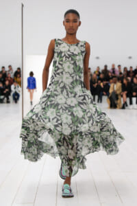 Model on a white runway wearing a sleeveless black floral print gown with a billowing, layered hem and turquoise shoes.