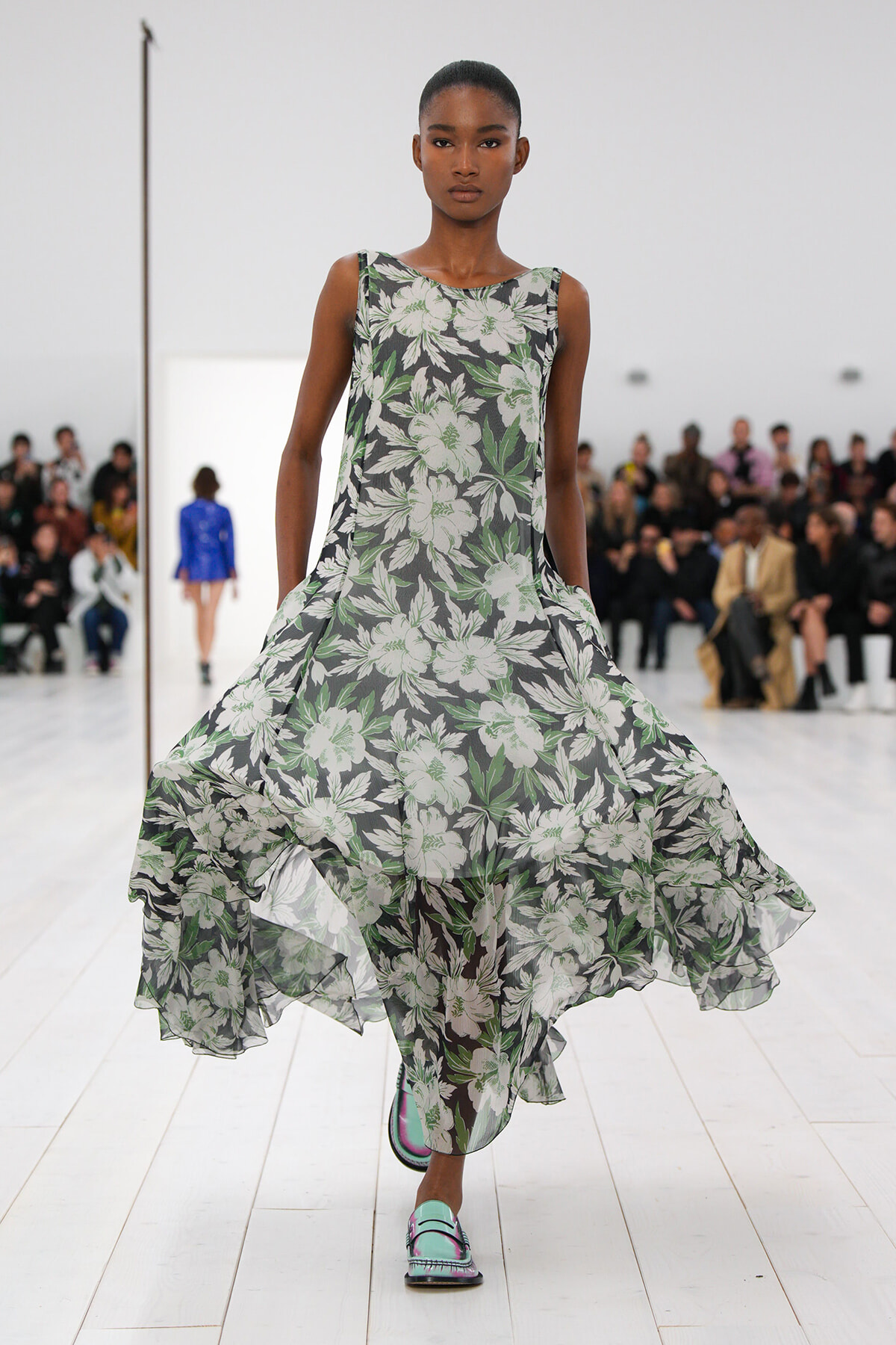 Model on a white runway wearing a sleeveless black floral print gown with a billowing, layered hem and turquoise shoes.