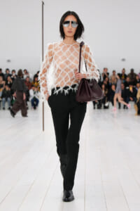 Model on a white-runway wearing a sheer feathered top, black tailored pants, dark mirrored sunglasses, and a burgundy handbag.