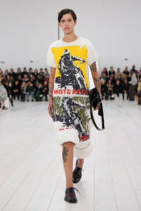 Runway model in a white graphic T-shirt with a bold yellow Moto Race print, camo-pattern pants with fur ankle cuffs, and brown lace-up shoes, holding a black handbag.