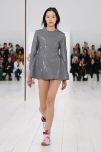 Model walking a runway in a grey sequined A-line mini dress with long sleeves, paired with pink and white sneakers.