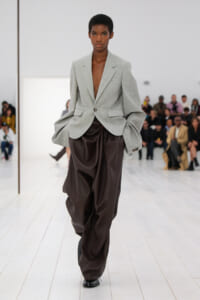 Model walks the runway in a light gray oversized blazer paired with dark brown draped satin pants, audience in the background.