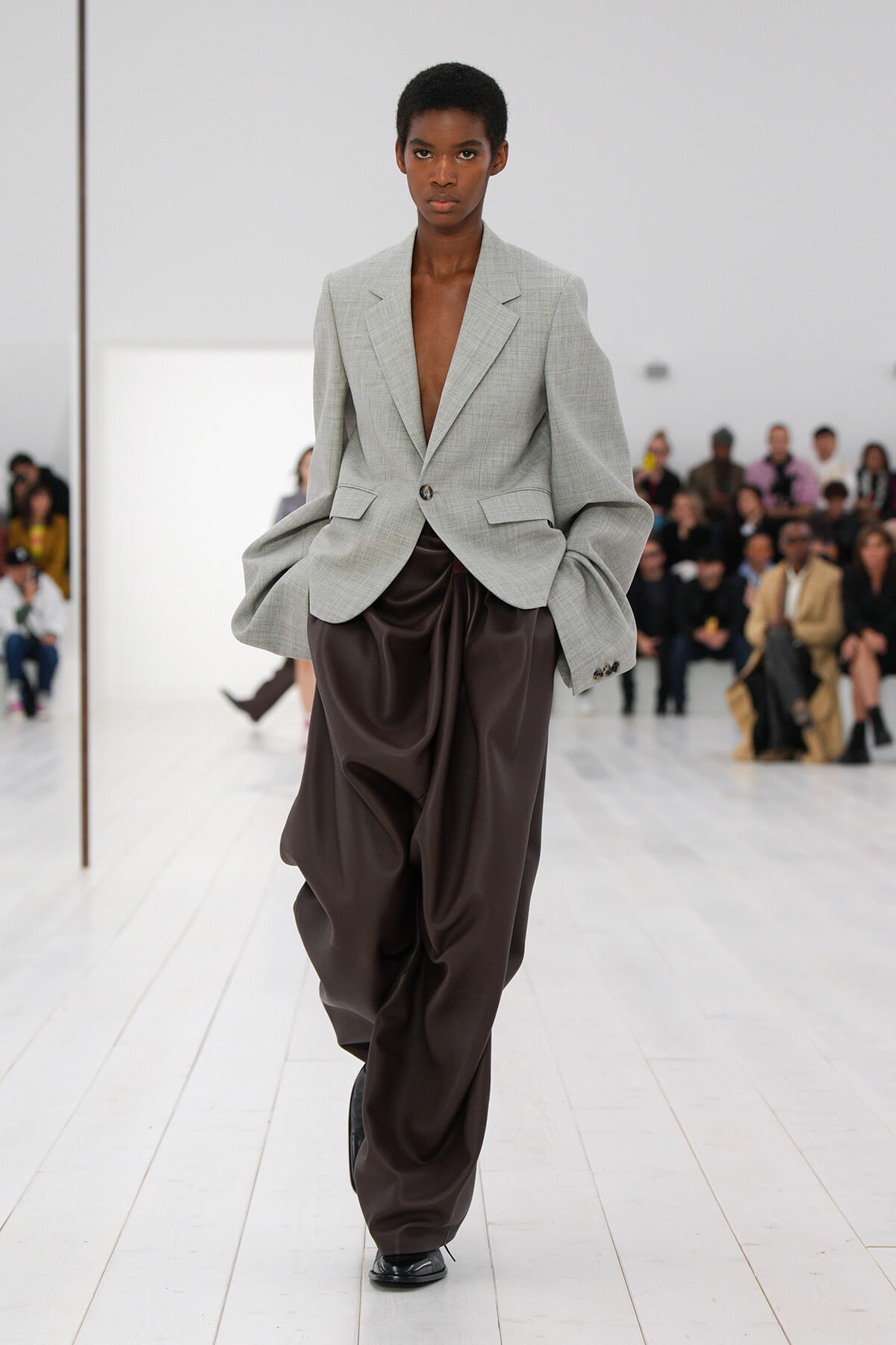 Model walks the runway in a light gray oversized blazer paired with dark brown draped satin pants, audience in the background.