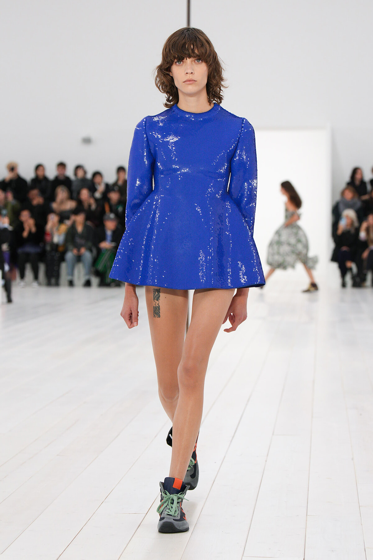 Model walking a runway in a bright royal blue sequined mini dress with long sleeves, wearing chunky sneakers.