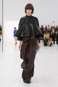 Model walking a runway in a black leather cape jacket with a diagonal zipper, paired with brown draped wide-leg pants and a Loewe belt.