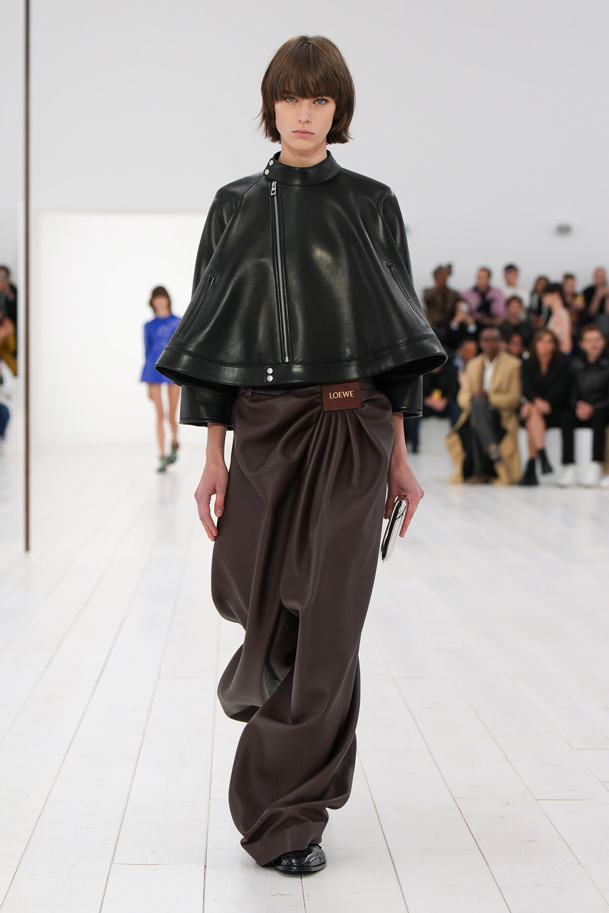 Model walking a runway in a black leather cape jacket with a diagonal zipper, paired with brown draped wide-leg pants and a Loewe belt.