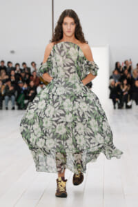 Model walking a runway in a sheer, floral-print maxi dress with off-shoulder, ruffled sleeves; audience in background.