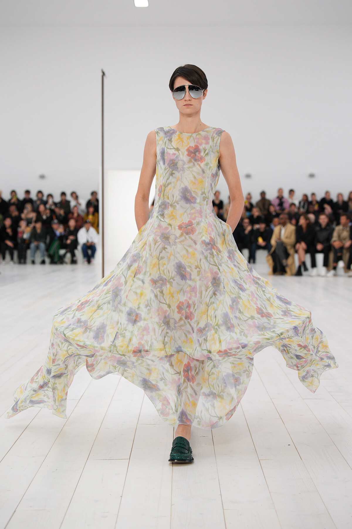 Model on a runway wearing a sleeveless, pastel floral maxi dress; dark sunglasses and green loafers, audience in the background.