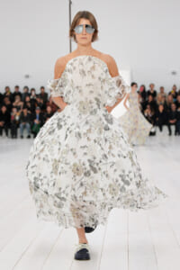 Model walking the runway in a white floral off-the-shoulder gown with voluminous, sheer skirt and ruffled sleeves, wearing mirrored sunglasses.