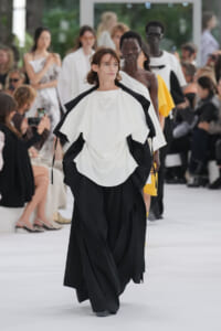 Model walks a fashion runway in a white oversized top with black layered accents; audience sits along a white catwalk behind her.