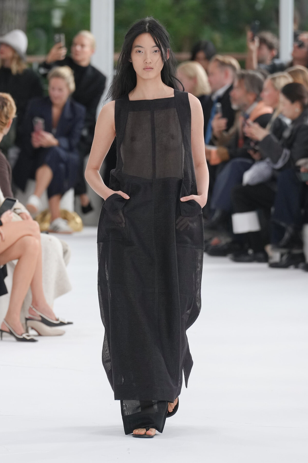 Model walking the runway in a sheer black, sleeveless maxi dress with pockets, open-toe sandals; audience in background.