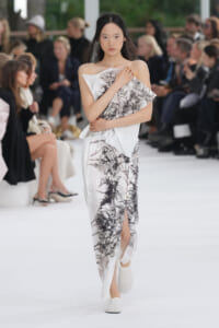 Model walking a runway in a white off-shoulder dress with a black forest print, hands crossed at the chest, audience in the background.