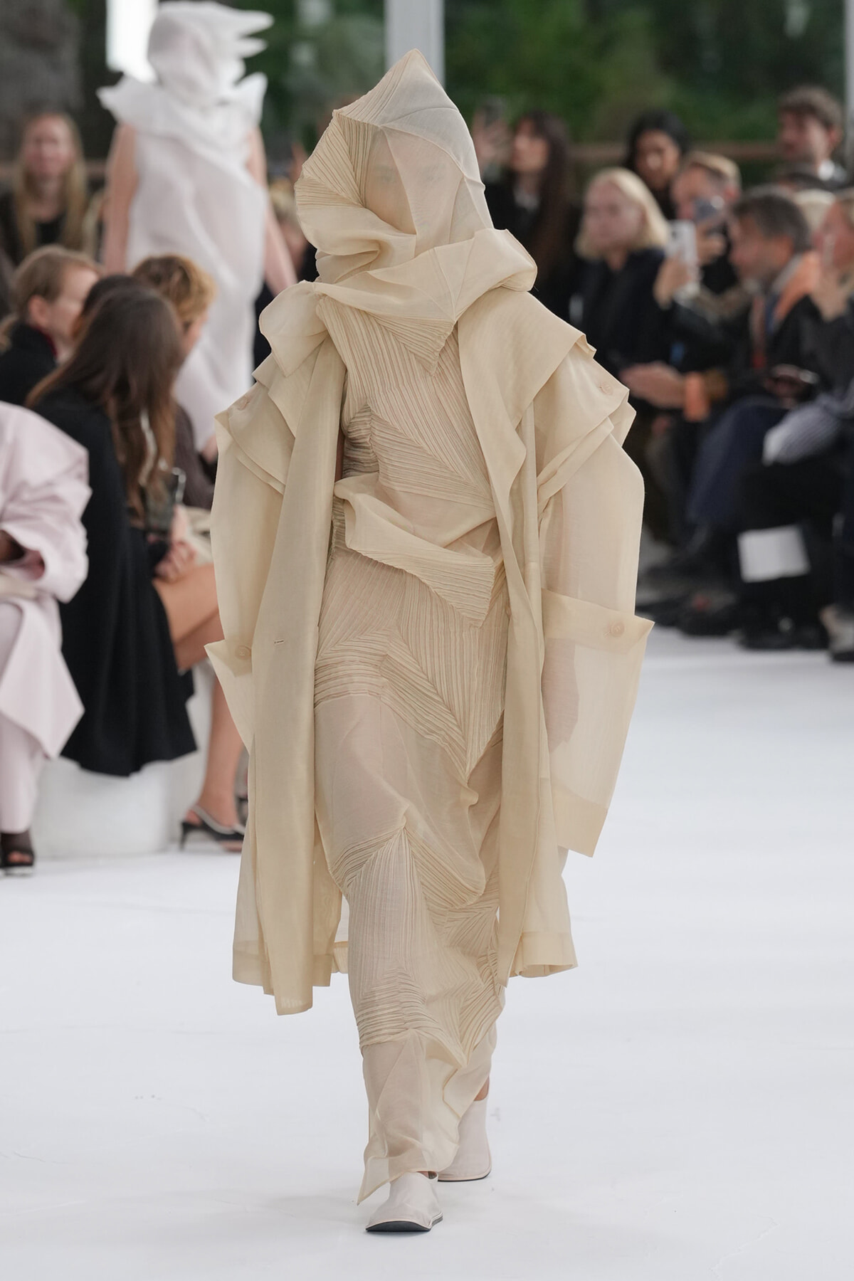 Model on runway wearing an off-white layered sculptural coat with a hooded headpiece.