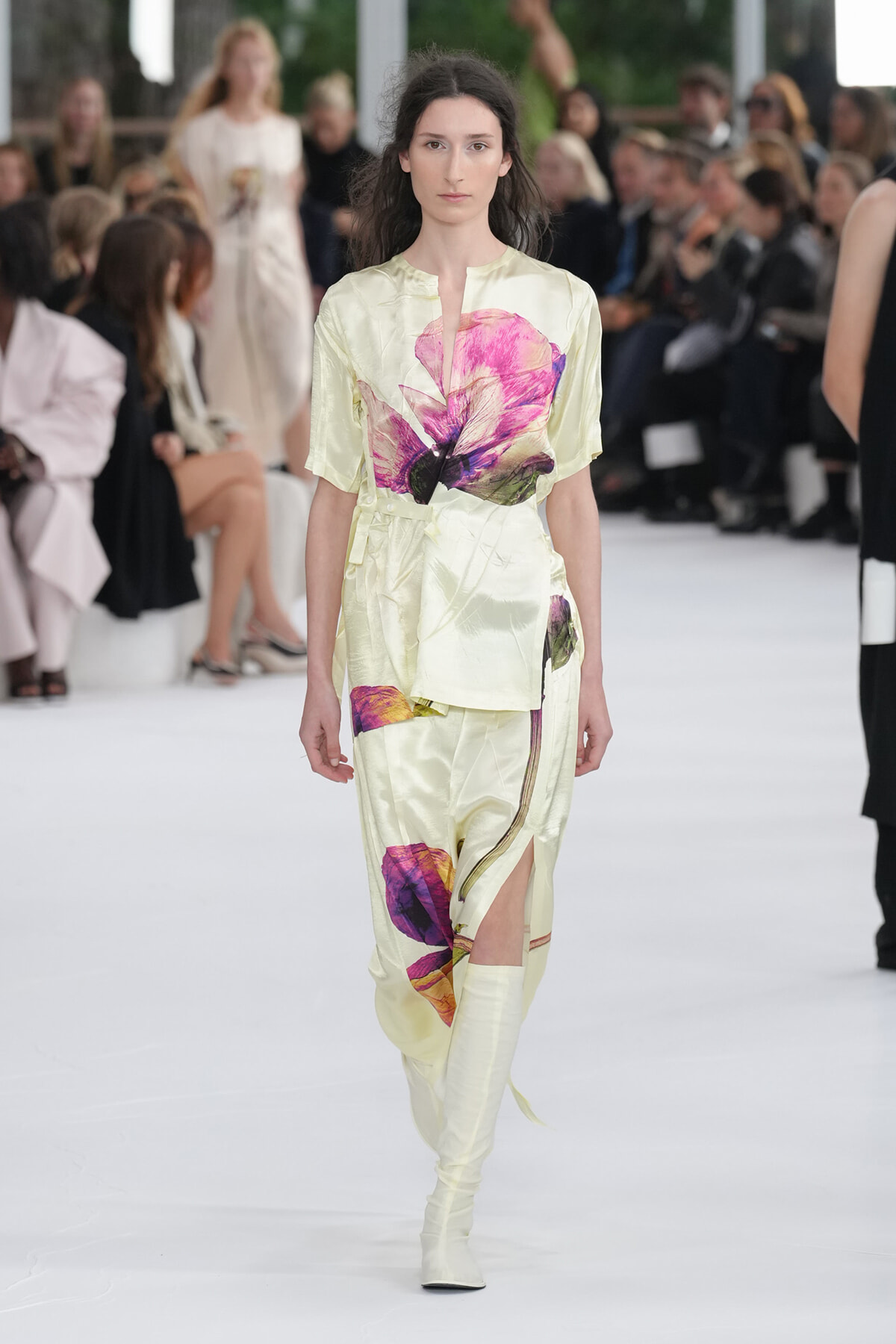 Model walks a pale yellow satin two-piece dress with large pink and purple floral print on a white runway; spectators blur in the background.