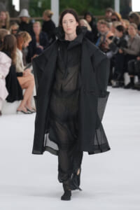 Model walks the runway in a black, oversized cape over a sheer, pleated dress with a high collar.