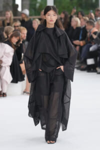 Model walks a black, sheer, layered cape-like outfit with wide trousers on a runway, audience watching in the background.