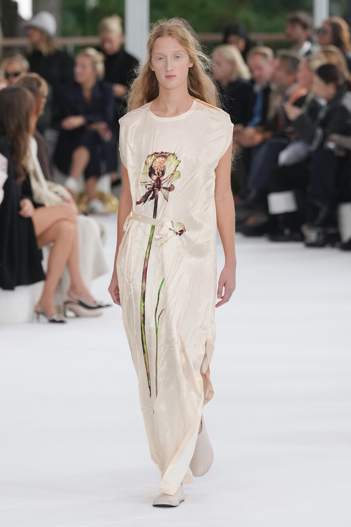 Model walking down a runway in a pale pink, ankle-length dress with a tall floral print stem design down the front.